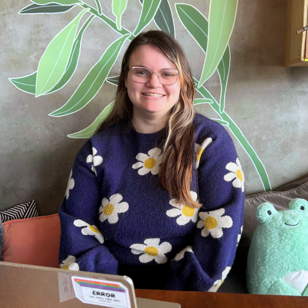 Person wearing a floral sweater sitting at a table with a laptop, in front of a wall with green leafy patterns.