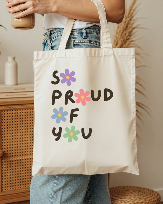 Person holding a tote bag with 'SO PROUD OF YOU' text and colourful flowers.